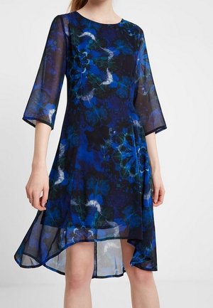 Robe florale légère de couleur bleue avec des manches 3/4, un ourlet asymétrique et un fond noir. Présente des motifs audacieux de bleu et de vert sur l'ensemble.