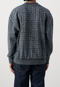 Sudadera gris con un estampado de logo tonal en toda la prenda, cuello redondo y puños y dobladillo acanalados. Combinada con pantalones de mezclilla oscuros.