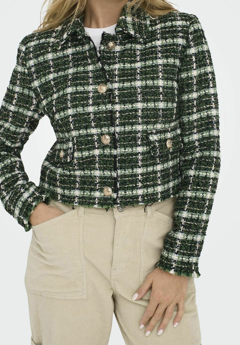 Femme portant une veste texturée à carreaux verts et blancs avec des boutons dorés et un pantalon en velours côtelé beige, les mains dans les poches et une posture détendue.