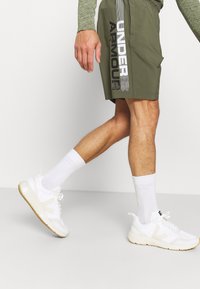 Olivgröna shorts med "Under Armour" textdesign, ihop med vita sneakers och vita kortsockor. Slätt tyg och avslappnad passform.