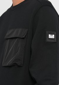 Black long-sleeve shirt made of cotton with a textured, slightly wrinkled patch pocket; features a logo patch on the sleeve.