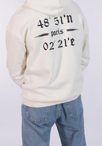 Sudadera color crema con texto negro en la parte de atrás, que presenta las coordenadas "48° 51'n" y "02° 21'e," combinada con jeans de color azul claro.