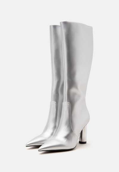 Paire de bottes montantes argentées brillantes avec bouts pointus et talons blocs cylindriques sur un fond clair uni.