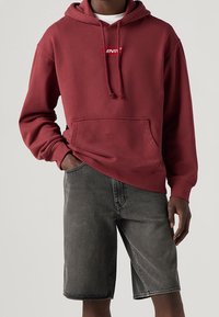 Rote Kapuzenjacke mit einer Vorderentasche und Logodetail, kombiniert mit grauen Denim-Shorts. Das Material wirkt weich und lässig.