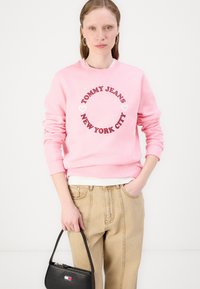 Rosa hettegenser med rund logo som har "TOMMY JEANS NEW YORK CITY" i fete, kastanjefargede bokstaver, sammen med beige bukser med høy midje og en svart håndveske.