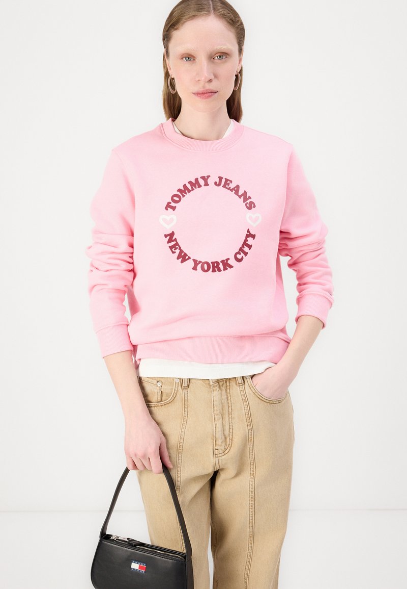 Felpa rosa con logo rotondo che presenta "TOMMY JEANS NEW YORK CITY" in grassetto color marrone scuro, abbinata a pantaloni/beige a vita alta e una borsa nera.