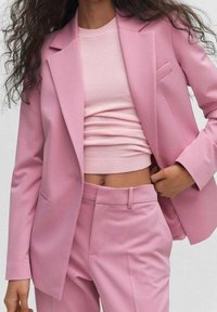 Femme portant un blazer rose cintré, un pantalon taille haute assorti et un haut en maille rose court, dévoilant le ventre, ajustant sa veste d'une main.