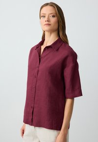 Camicia in lino borgogna con maniche corte, chiusura a bottoni sul davanti e vestibilità rilassata. Il tessuto ha un aspetto testurizzato e leggermente stropicciato.