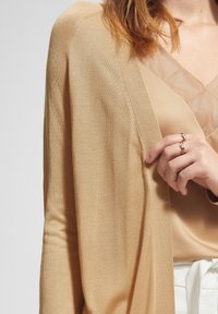 Beige cardigans med ett lätt, texturerat tyg, bärs över en tan-färgad topp. Framträdande V-halsringning och en hand som håller en vågig ring.