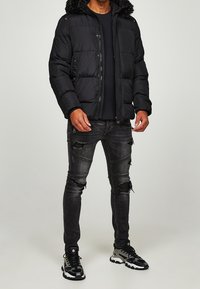 Muž oblečený v černé péřové bundě, roztrhaných černých úzkých džínách a černošedých teniskách, stojící před jednobarevným světlým pozadím.