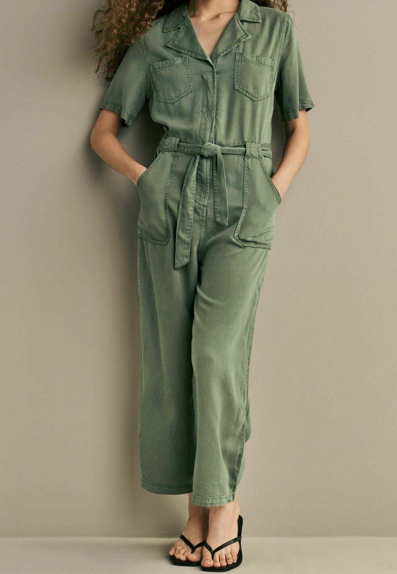 Femme debout portant une combinaison ceinturée vert olive à manches courtes et des sandales à talons noires sur fond beige.