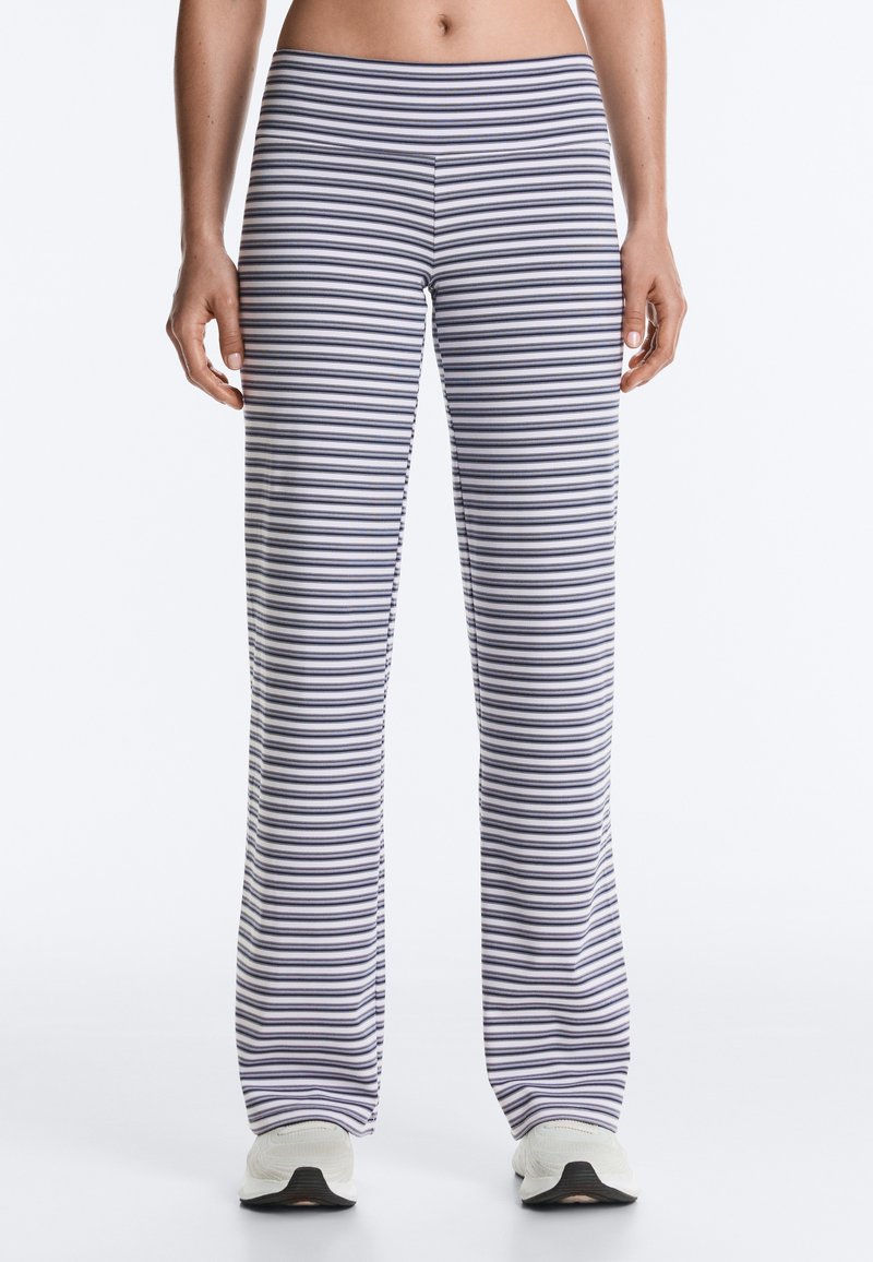 Le bas du torse et les jambes d'une femme portant un pantalon blanc à rayures noires horizontales et des baskets blanches, debout devant un fond uni.