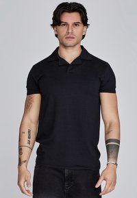 Melna īsām piedurknēm polo krekls ar V veida kakla izgriezumu un platām apkaklēm. Mīksta, ribota tekstūra. Modelis valkā tumšas džinsas, redzami roku tetovējumi.