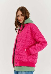 Prešívaná ružová bunda so striebornými cvokmi, pod ktorou je zelená mikina s kapucňou, umiestnená na neutrálnej pozadí.