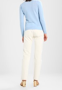 Suéter azul claro de tejido suave, combinado con pantalones blancos. Presenta una silueta ajustada, mangas largas y sandalias de tacón de madera con espalda abierta.