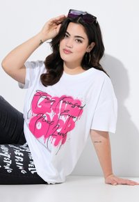 Camiseta blanca de gran tamaño con texto estilo graffiti en rosa y negro. El modelo lleva vaqueros negros con letras blancas y gafas de sol moradas.
