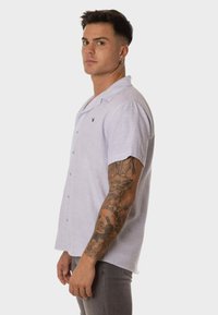 Camisa de botones de manga corta en un suave color lavanda, confeccionada en un tejido texturizado, que presenta un cuello con muesca, botones en la parte delantera y un logo en el lado izquierdo del pecho.