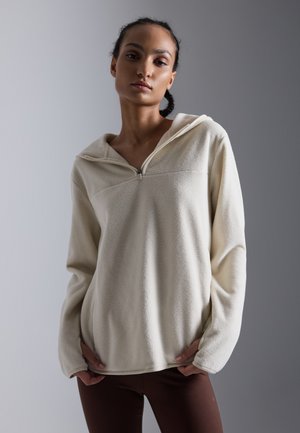 Kremfarget fleece-pullover med glidelåshette, med teksturert overflate, lange ermer og en avslappet passform. Kontraststikning synlig.