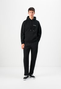Young man wearing a black hoodie, black pants, and black sneakers with white laces standing against a plain white background.