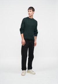 Σκούρο πράσινο πουλόβερ με πλέξη καλωδίων και στρογγυλή λαιμόκοψη, συνδυασμένο με μαύρα τζιν και λευκά αθλητικά παπούτσια. Το ύφασμα έχει υφή και σχέδιο.