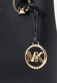 Sort tekstureret lædertaske med et guldfarvet cirkulært logocharms med "MK" og en firkantet nitte.