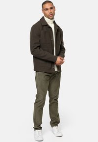 Chaqueta de lana marrón con dos bolsillos frontales, cierre de botones, llevada sobre un suéter de cuello alto color crema, combinada con pantalones verdes oliva y zapatillas blancas.