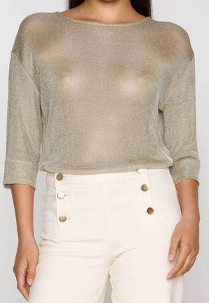 Vrouw draagt een lichtgouden, doorzichtig, fijngebreid shirt met lange mouwen en een witte, hoge taille broek met zes gouden knopen aan de voorkant.