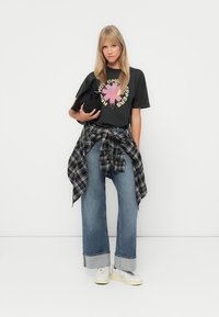 Black graphic t-shirt with colorful print, loose light blue jeans with rolled cuffs, flannel shirt tied at waist, and white sneakers.