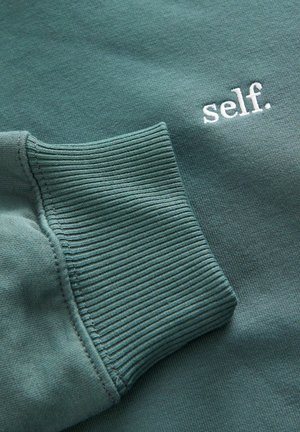 Zeleni pamučni dukserica sa rebrastim manžetama i izvezenim natpisom "self." u bijeloj boji. Meka tekstura s ležernim dizajnom.