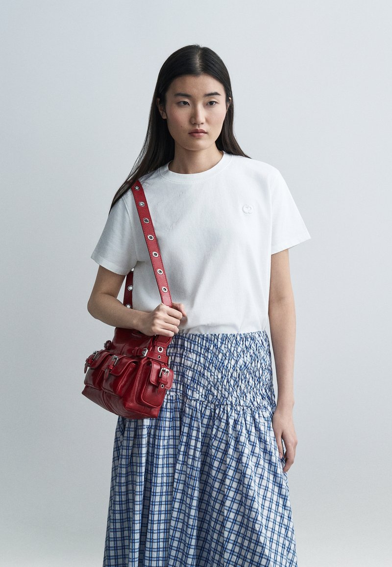 Femme portant un t-shirt blanc et une jupe à carreaux bleue, tenant un sac à bandoulière en cuir rouge avec plusieurs poches.