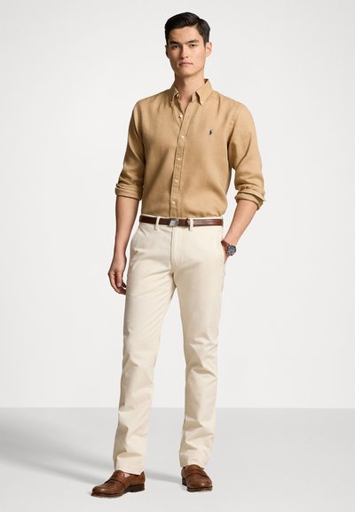 Camicia button-up marrone chiaro con maniche arrotolate, abbinata a pantaloni beige e scarpe marroni. L'outfit presenta una fibbia in argento e un orologio.
