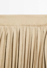 Beige pleadisella kangas, jossa on pehmeä tekstuuri, pystysuorat poimut ja tasainen yläreuna. Materiaalissa on hieno kiilto ja luonnonkuituja.