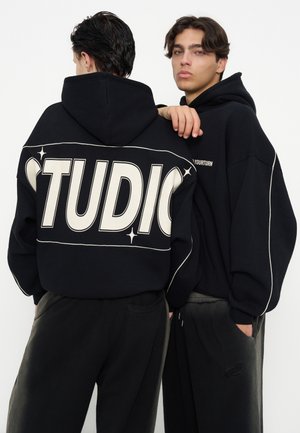 Zwei junge Männer in schwarzen Kapuzenpullovern und Hosen posieren vor weißem Hintergrund, einer sieht nach vorne, der andere zeigt die Rückseite des Kapuzenpullovers mit dem Text "STUDIO".