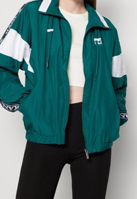 Turkusowa kurtka przeciwdeszczowa z białymi paskami, z zamkiem z przodu i elastycznymi mankietami. Posiada kaptur ze sznurkiem i szczegóły z logo na rękawach.