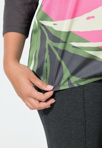 Värikäs pusero, jossa on vaaleanpunainen ja vihreä lehtikuvio, lyhyet mustat hihat ja sileä pinta. Alla näkyy harmaat, vartalonmyötäiset legginsit.