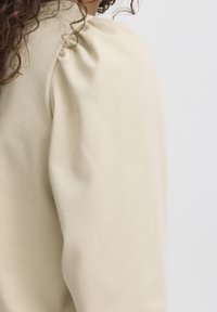 Hellbeiger Pullover mit puffierten Schultern, weichem Textil und glatter Oberflächenstruktur. Die Seitenansicht zeigt die Detailansicht der Ärmel.