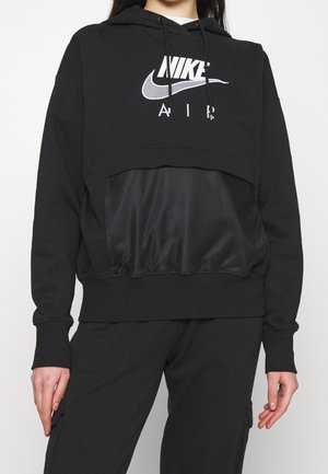 Persoon draagt een zwarte Nike Air hoodie met een mesh voorvak en bijpassende zwarte broek tegen een effen achtergrond.