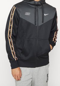 Svart Nike zip-up hoodie med grå detaljer, som har orange och vita sidränder, en huva och framfickor. Strukturerad tyg.