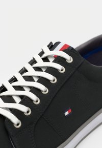 Zapatilla de lona negra con cordones blancos, que presenta un pequeño logo bordado en rojo, blanco y azul, y una suela de goma gris.
