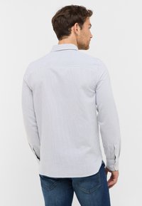 Biała koszula w niebieskie pionowe paski, z długimi rękawami, z zapięciem na guziki przy mankietach oraz z yoke z tyłu. Tkanina ma gładką fakturę i wyrazisty wygląd.