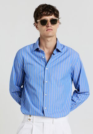 Mann mit durchsichtigen Sonnenbrillen, blau gestreiftem Hemd und weißen Hosen, der vor einem einfarbigen hellen Hintergrund steht.