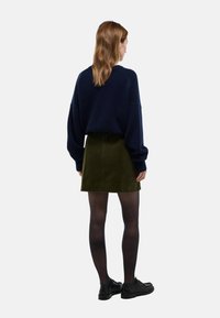 Femme aux longs cheveux bruns portant un pull bleu marine, une jupe vert olive, des collants noirs et des chaussures noires, debout de dos sur un fond blanc.