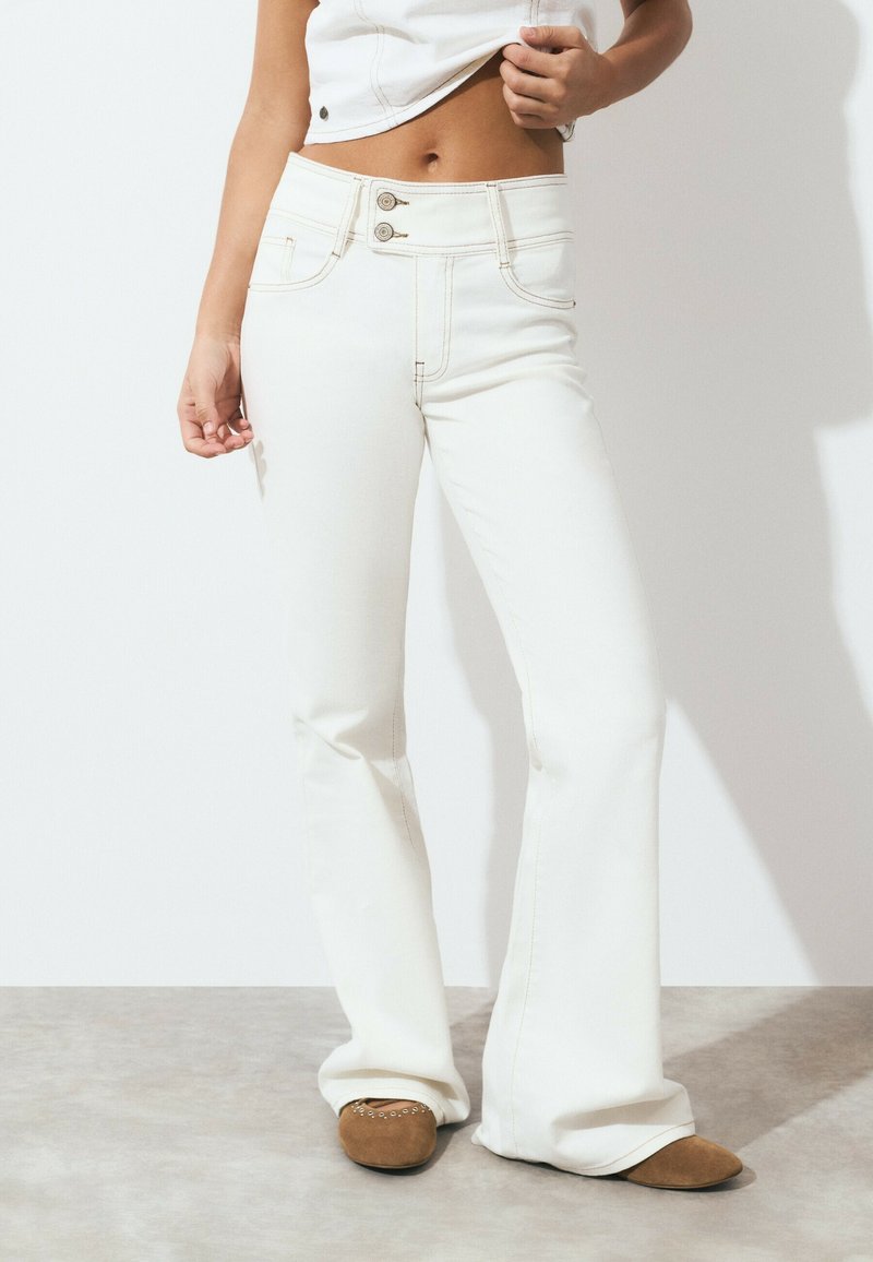 Woman wearing high-waist white jeans with double button closure and brown suede pointed flats, standing on gray floor against white wall.