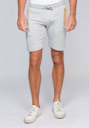 Homme portant un short gris clair avec cordon, poches zippées jaunes et baskets blanches, debout sur un fond clair.