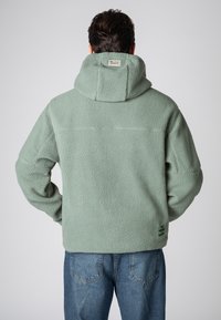 Fleece jas in lichtgroen, met een textuur aan de buitenzijde, een capuchon en zichtbare stiksels. Etiket op de achterkant met de merknaam.