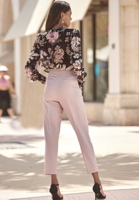Blusa floreale in tonalità scure, tessuto trasparente con maniche a sbuffo, abbinata a pantaloni rosa chiaro, vestibilità su misura, e sandali neri con tacco.