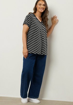 Zwart-wit gestreept V-hals T-shirt gecombineerd met hoge blauwe denim broek en witte sneakers, staande tegen een lichte muur.