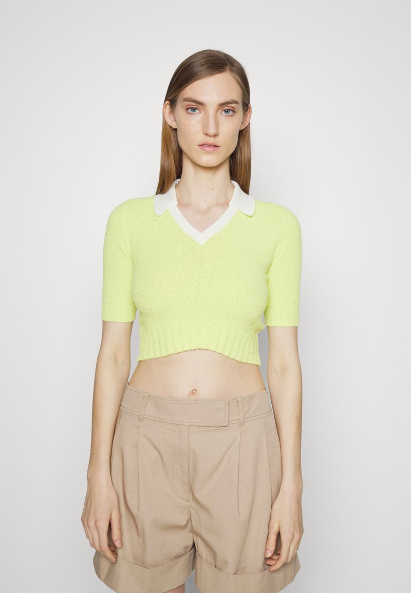 Sweater cropped de manga curta em verde limão brilhante com um decote de colarinho branco. Tecido texturizado com uma bainha canelada. Combinado com calções bege.