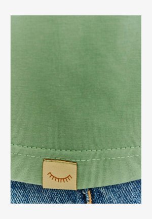 Svetlozelená bavlnená košeľa s lemom a béžovou nášivkou s jednoduchým dizajnom oka, skombinovaná s modrými rifľami. Hladká textúra a čisté švy.