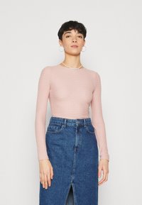 Rosa Langarmoberteil mit strukturiertem Muster, kombiniert mit einem blauen Jeansrock mit Schlitz vorne. Schlichte Schmuckstücke runden den Look ab.